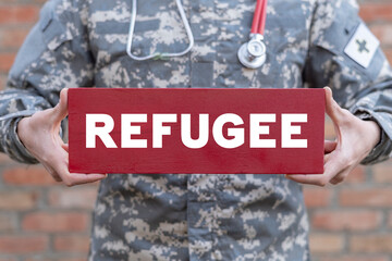 Military medic holding wooden card with refugee word. Concept of refugee medical assistance. Safe...