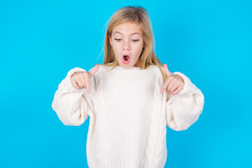 Amazed caucasian little kid girl wearing wool jersey over blue background points down with fore...