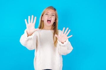 Dissatisfied caucasian little kid girl wearing wool jersey over blue background frowns face, has disgusting expression, shows tongue, expresses non compliance, irritated with somebody. - obrazy, fototapety, plakaty
