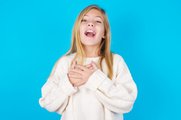 Happy smiling caucasian little kid girl wearing wool jersey over blue background has hands on chest...