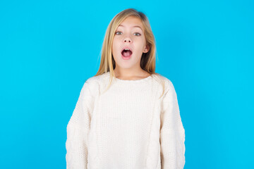 Emotional attractive caucasian little kid girl wearing wool jersey over blue wall with opened mouth...