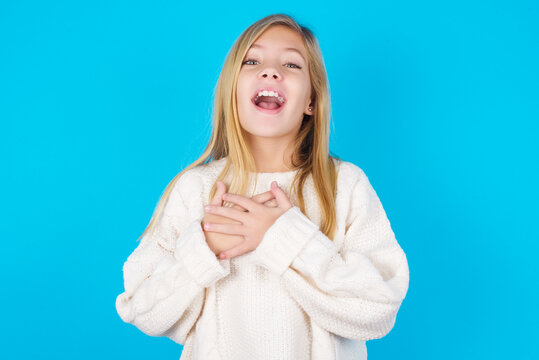 Happy smiling caucasian little kid girl wearing wool jersey over blue background has hands on chest near heart. Human emotions, real feelings and facial expression concept.