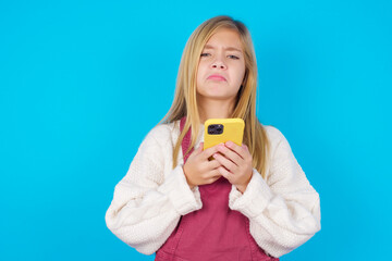 Portrait of a confused caucasian little kid girl wearing jumpsuit over blue background holding mobile phone and shrugging shoulders and frowning face.