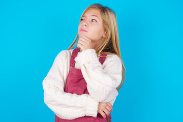 Thoughtful caucasian little kid girl wearing jumpsuit over blue background holds chin and looks away pensively makes up great plan