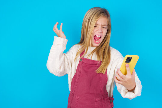 Photo of outraged annoyed caucasian little kid girl wearing jumpsuit over blue background holds cell phone, makes call, argues with colleague,  expresses negative emotions. People and anger.
