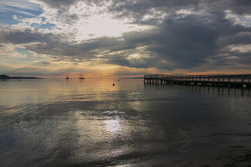 Fototapeta premium Beach scene with silhouette of pier and sailing vessels in the bay.