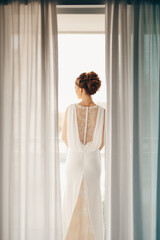Bride in a white lace dress with a beautiful high hairdo stands on the balcony. View from the room
