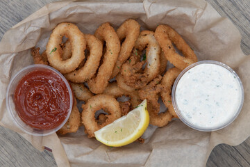 Overhead view of crispy fried calamari rings served in a paper lined basket with dipping and tarter sauce for a crunchy meal