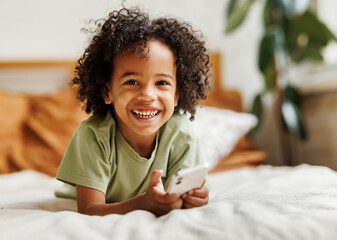 Cute little african american boy with smartphone watching cartoons or playing mobile games at home