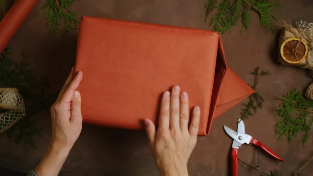 Christmas preparation, DIY holiday presents packaging concept. Female hands wrapping Christmas gift box with red kraft paper. Overhead view, POV. 