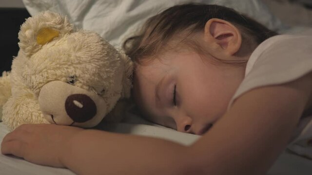 Child, daughter sleeps and hugs her favorite teddy bear toy. The kid sleeps at home on the sofa in the nursery. Sleeping healthy baby in bed next to a teddy bear. Childhood happiness concept.
