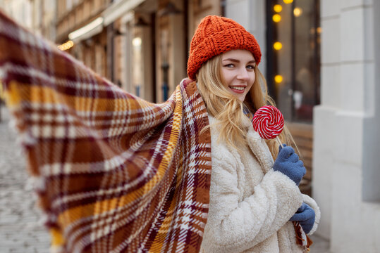 Happy smiling woman holding lollipop, posing in street. Lady wearing stylish knitted beanie hat, checkered scarf, faux fur coat. Copy, empty space for text