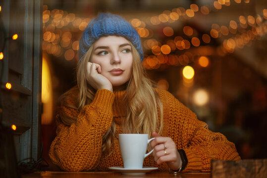 Cute thoughtful woman holding cup of coffee or tea, sitting, posing at cafe. Winter vacation, Christmas holidays conception. Model wearing knitted beanie hat, sweater. Copy, empty space for text