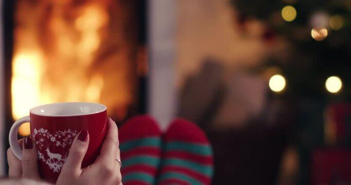 Over the shoulder young woman relaxing near fireplace with cup of tee in cozy christmas woolen socks with decorated xmas tree in background shot in 4k super slow motion