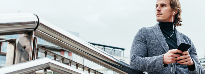 Modern businessman with smartphone on a city street. Handsome man wearing earphones and speaking on mobile phone, listening music or podcast outdoors.