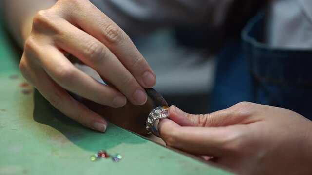 Close-up gems on table in workshop with female hands engraving stones in silver ring in slow motion. Unrecognizable expert skilled Caucasian woman working in jewellery workshop indoors