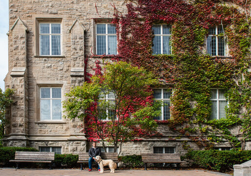 Man and dog sitting in front of red ivy covered building in fall.