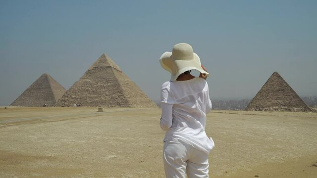 Tourist woman with white straw hat in front of The Great Giza pyramids in Cairo, Egypt enjoying the view of the Egyptian and world UNESCO ancient heritage while on summer vacation