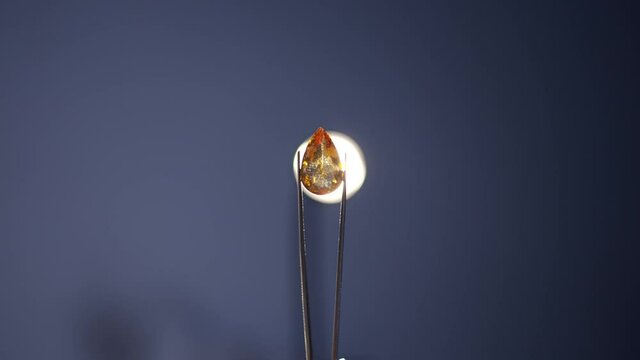 Close-up shiny orange precious gemstone in tweezers with lamp at background. Brilliant luxurious high-quality fine gem in light in workshop indoors. Slow motion