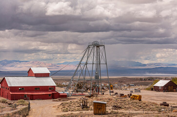 Tonopah, Nevada, US - May 18, 2011: Historic Mining Park. Red building adjacent to gray metal shaft tower with rotar on sandy desert floor under heavy gray cloudscape. Mountain range in back, - Powered by Adobe