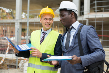 European man engineer with laptop and African-american foreman with documentation standing on building site and discussing construction.