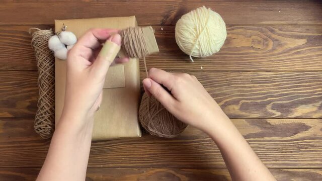 Person's hands wind beige yarn on piece of cardboard. Handmade crafting decorations of gift for Christmas or Birthday. Box in craft wrapper, twine, cotton and scissors on wooden table. Festive mood.
