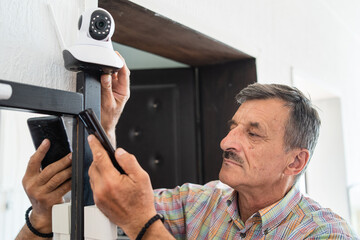 Caucasian senior man holding home security surveillance camera and mobile phone trying to install...