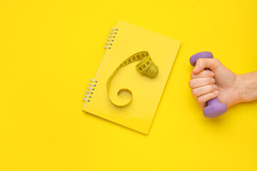 Female hand with dumbbell, notebook and measuring tape on yellow background