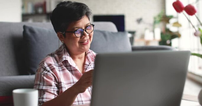 Mature Asian woman in checkered shirt entering credit card credentials into netbook and celebrating success while sitting on sofa and making online order in living room at home
