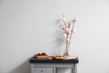 Glass vase with cotton flowers on table near light wall