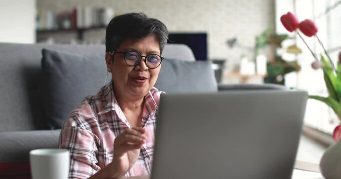 Positive mature Asian woman talking and gesturing thumb up while making video call via netbook during remote work from home