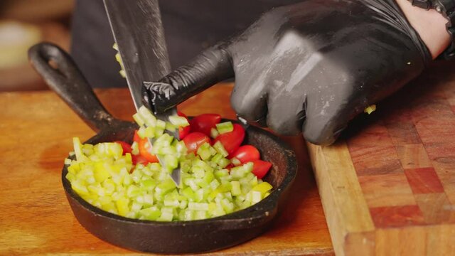 Preparation of salad ingredients, professional chef wearing gloves, transferring diced green bell pepper into stone pan next to juicy cherry tomato.