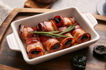 Baking dish with delicious prunes baked in crispy bacon and rosemary branch on wooden board