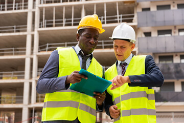 Two civil engineers working at the construction site are discussing the construction plan, holding documents in their hands and carefully studying them