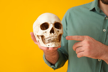 Handsome man pointing at human skull on yellow background