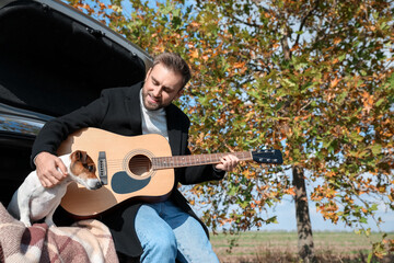 Handsome man with cute dog playing guitar in car trunk on autumn day