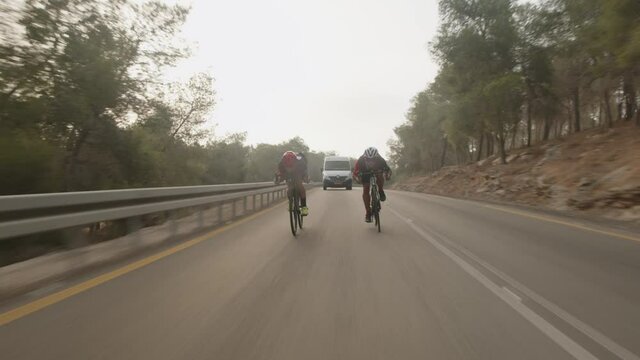 two road cycelist professionals riding in the desert while a big white van behind them.