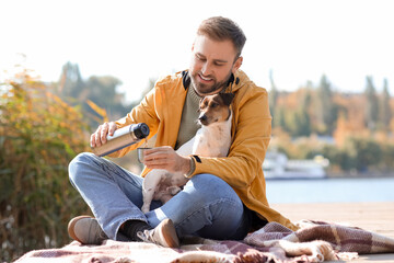Handsome man with cute dog pouring tea into cup near river on autumn day