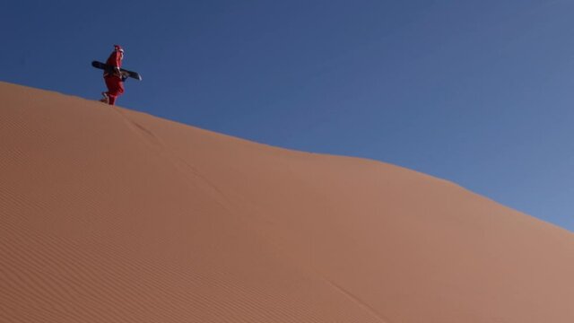Santa Claus Walking on the Ridge of A Huge Sand Dune with a Snowboard on a Sunny Day at the Desert
