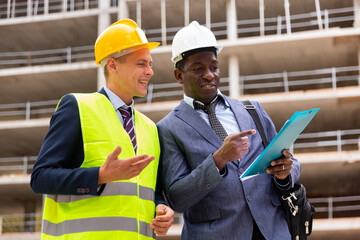 European foreman and African-american engineer in hardhats standing on building site and talking about documentation.