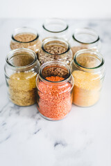 healthy whole grains and legumes in clear pantry jars on marble background including quinoa rice buckwheat lentils and barley, simple ingredients concept