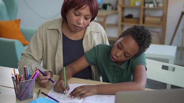 Family education with parents. Attentive little african-american schoolboy colors pictures spbd in notebook under mother control at workplace in living room