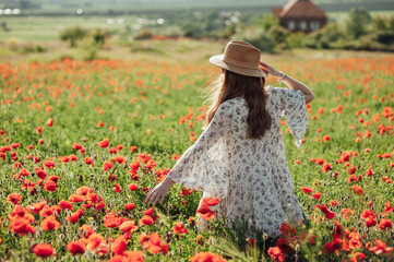 Girl wearing white dress and straw hat walks among red poppy flowers on the sunset.