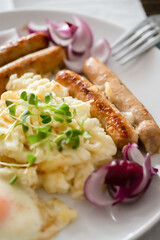 Pork sausages with onions, eggs, mashed potatoes and microgreens. Hearty breakfast. Dark wooden background, white ceramic plate. Top view.