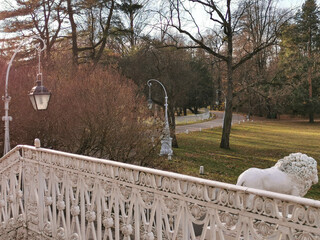 A white sculpture of a lion outside the fence of the Elaginoostrovsky Palace against the background of a beautiful lantern, trees and a sky with clouds