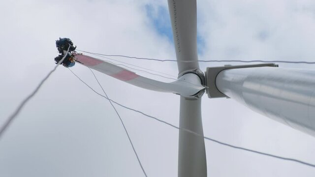 Service workers fix wind-driven generator blade sitting on climber seats with safety harnesses under sky with white clouds low angle shot