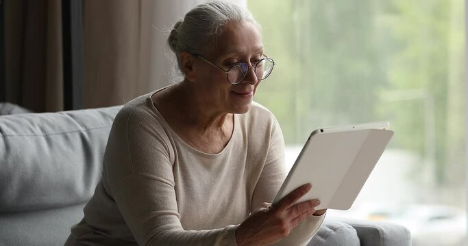 Smiling older age female in glasses rest on couch at home using digital tablet select electronic book at online library in reader app. Happy retired lady aged 60 years networking on touchpad device
