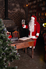 Santa Class holds candles in his hands for the New Year and Christmas 2022 at a large table against the background of a Christmas tree