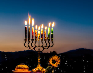 Fototapeta premium Close-up of festive burning wax candles that are traditional symbols of Hanukkah Holiday of Light, selective focus on burning candles and sunflower, blurred background with night mountains and sky 
