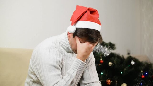 Loneliness in Christmas celebration. Sad, angry, young man in Santa hat in depressed mood sitting alone at home next to fallen decorated Christmas tree and aggressively hitting toys